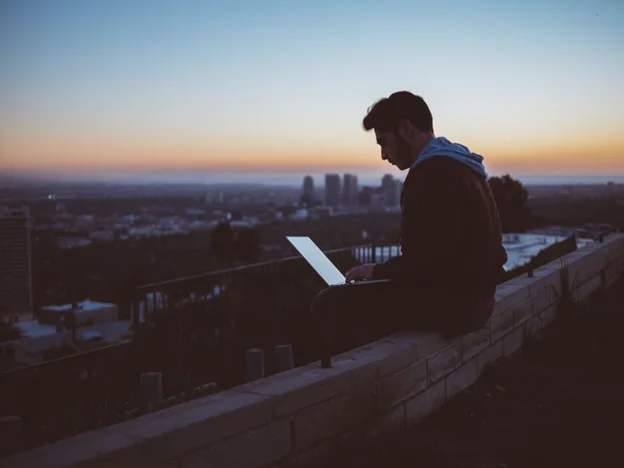 A man sits on a stone ledge at sunset, working on a laptop.